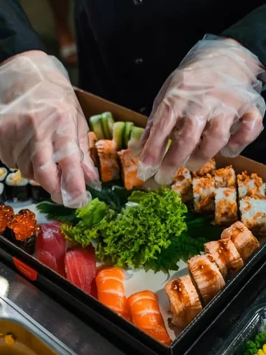 Zwei behandschuhte Hände richten eine Platte mit Nigiri, Maki und Inside-out-Rollen an